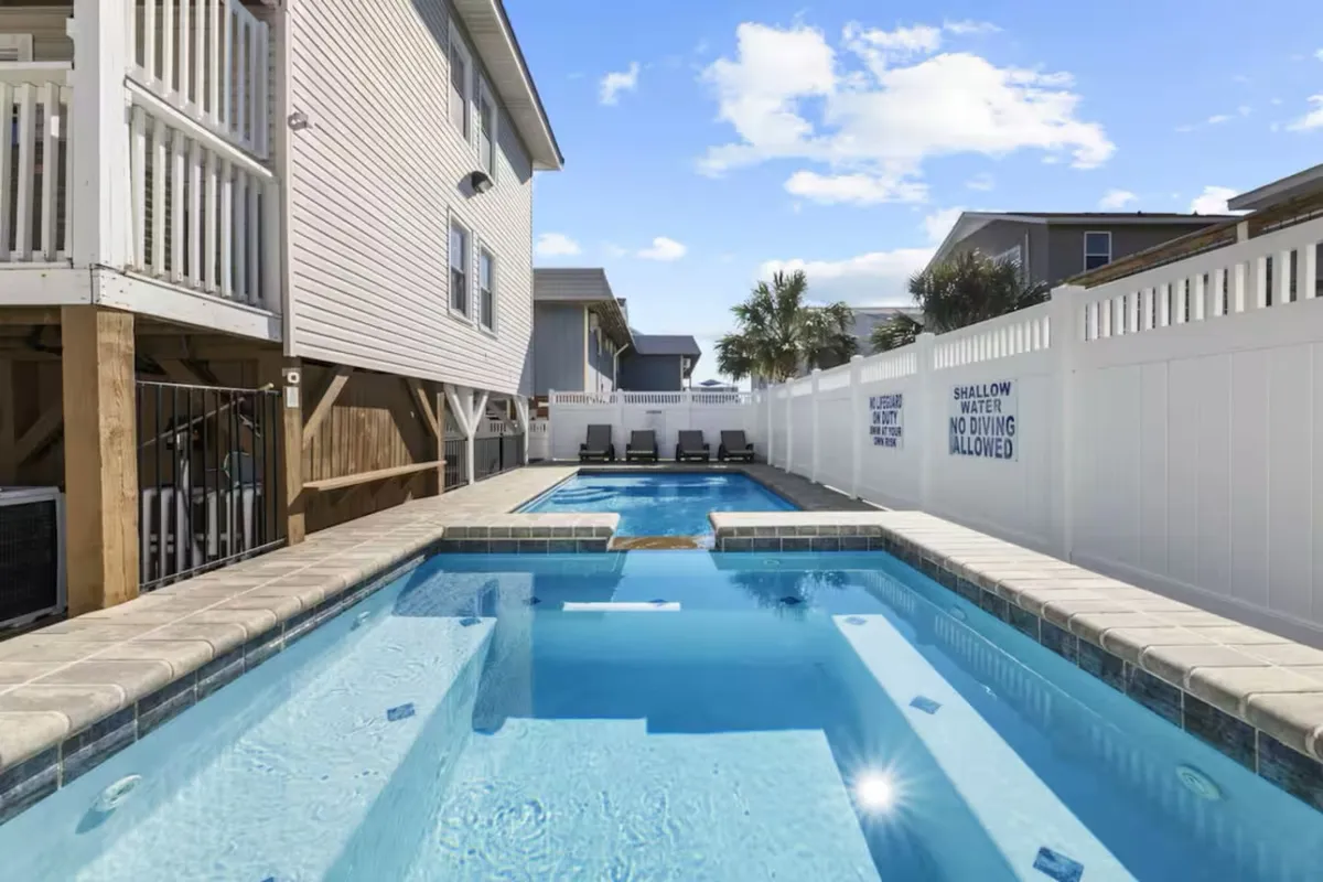 A bright exterior shot of a private swimming pool surrounded by lounge chairs and a secure fence, with neighboring buildings visible in the background. The setting suggests both daytime and evening relaxation, with space for swimming, sunbathing, and socializing.