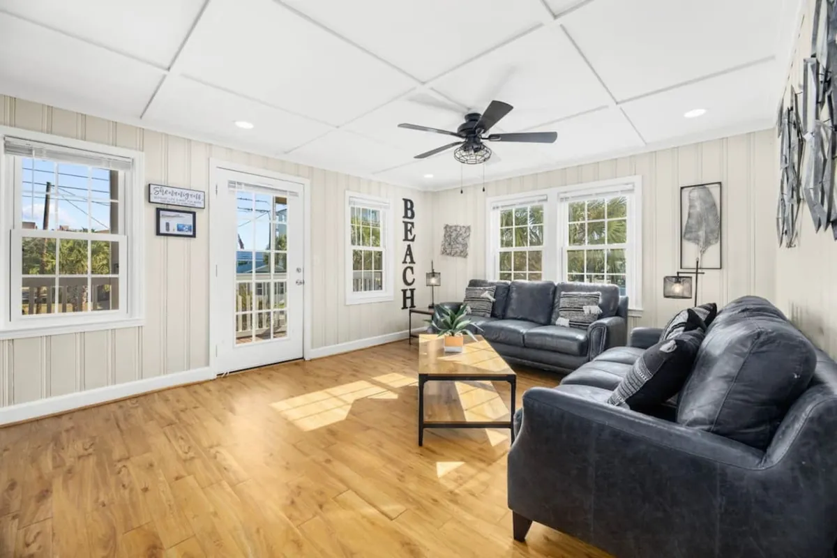A bright and cheerful living room with a plush sofa, a coffee table, decorative wall art including a playful 'BEACH' sign, and large windows letting in natural light. The room exudes warmth and invites group interaction or quiet relaxation.