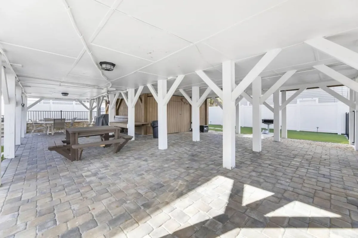 An outdoor dining area featuring a sturdy wooden picnic table set on stone flooring, shaded by support beams, and an adjacent outdoor kitchenette. The friendly, casual setup is ideal for al fresco meals, barbecues, and festive gatherings.