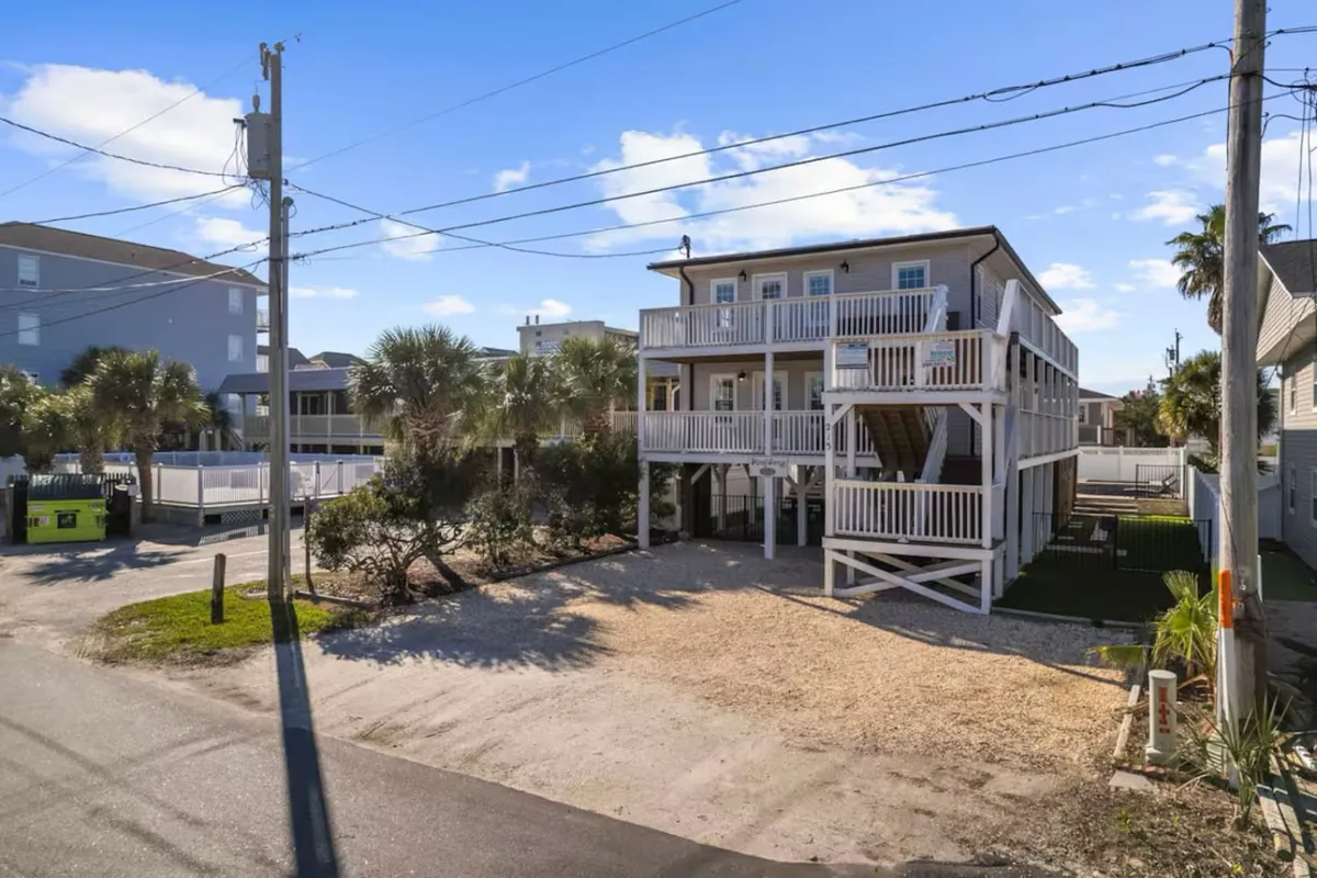 A welcoming two-story vacation home framed by palm trees, with ample open parking and a sunny front porch. The image feels inviting and hints at easy access to both the house and surrounding coastal attractions.
