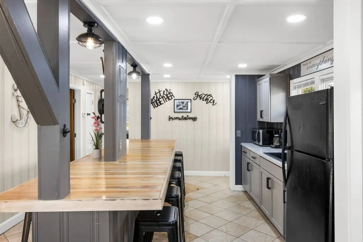 A kitchen scene centered around a kitchen island with a wood countertop and black bar stools, accented by decorative wall signs. The atmosphere is inviting and relaxed, with clear indications of space for group meal prep and enjoyment.