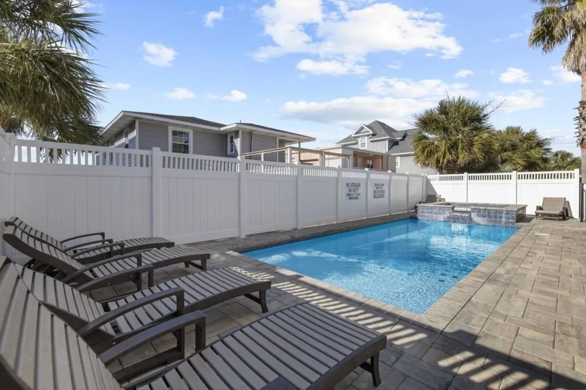 This image features a tranquil backyard scene centered on a sparkling swimming pool surrounded by lounge chairs, palm trees, and a secure fence. The pool area is inviting, with ample space for sunbathing, swimming, and children playing in the water. The atmosphere is serene, evoking the spirit of a private resort.