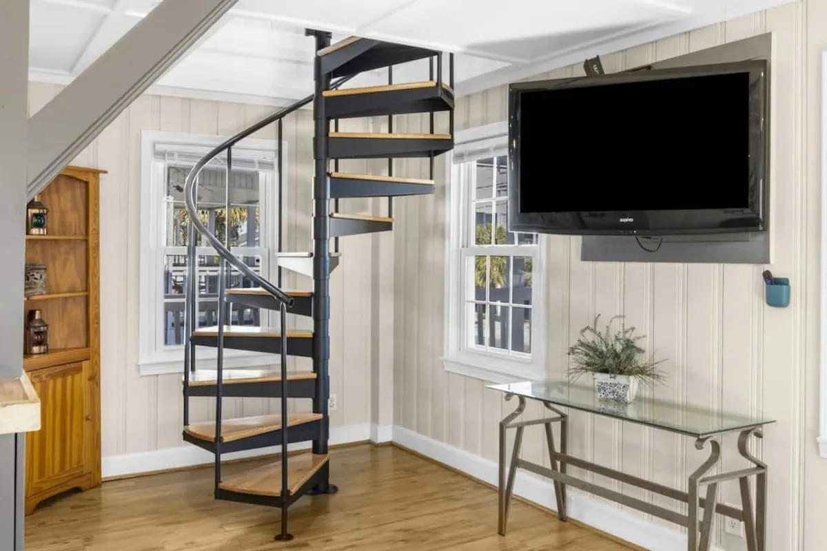 This image captures a bright, contemporary living area featuring a spiral staircase, a large TV mounted on the wall, a glass table with decorative accents, and a cozy wooden cabinet. The space is designed for relaxation and socializing, ideal for family gatherings or group activities.