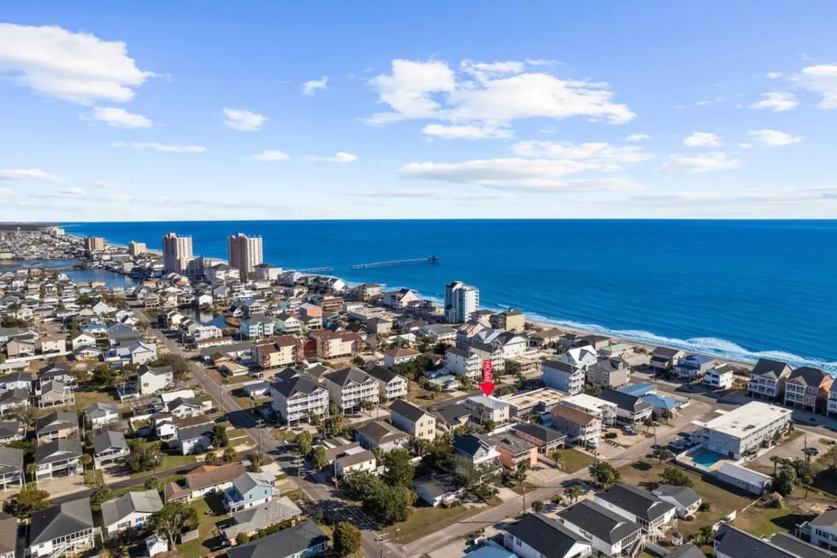 This image showcases a sweeping view of the ocean, the beachfront, nearby residential buildings, and the lively downtown area. The scene captures a mix of beach activity, water sports, and the vibrant atmosphere of local shops and dining establishments along the coast.