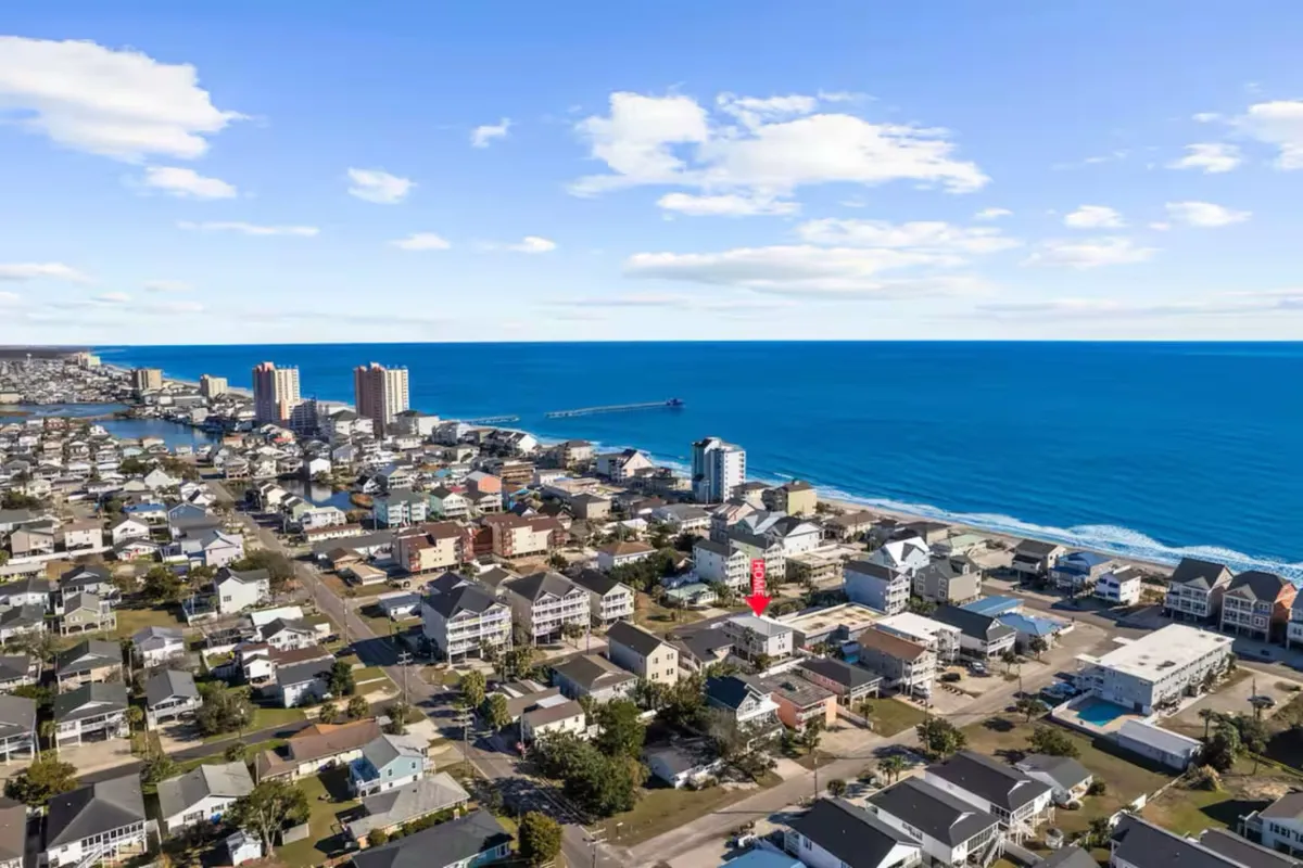 The image shows a spacious single-family house in North Myrtle Beach, set in a welcoming residential neighborhood with a vibrant view of the ocean at sunset. Surrounding the home are inviting outdoor spaces, while the proximity to both the shoreline and local shops is evident. The warm, relaxed atmosphere sets the stage for gatherings and beachside fun.