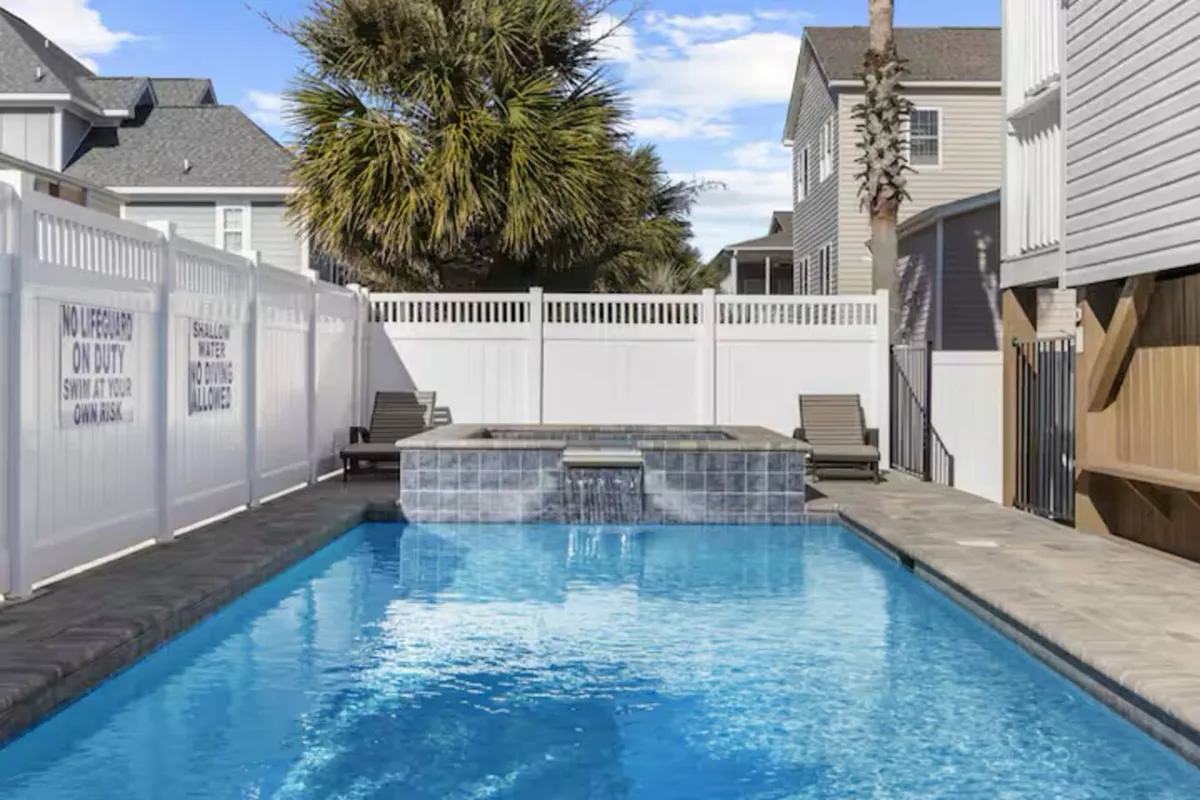 A leisurely backyard view showing both a shimmering swimming pool and a bubbling hot tub, accented by palm trees and secure fencing. The scene suggests relaxation and group enjoyment, with space for sunbathing, swimming, and soaking in the hot tub.