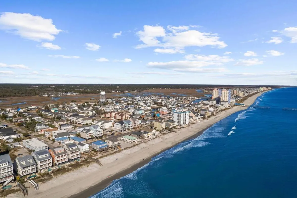 This image features a sweeping view of the ocean and sandy beach with the townscape and bright sky in the distance. The scene captures the vibrant shoreline, ocean waves, and the sense of possibility for water activities and relaxation along the coast.