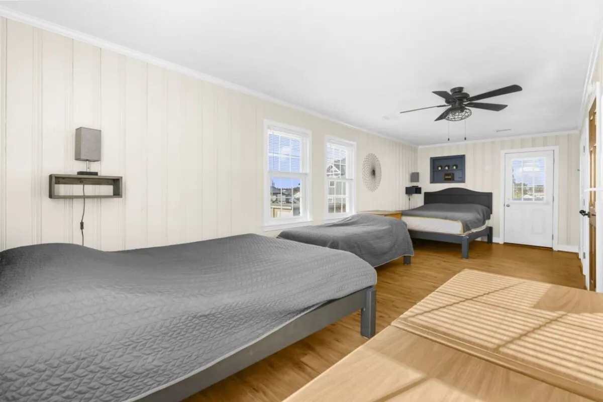 A spacious bedroom with two large beds, wooden flooring, and natural light streaming in from windows. The space feels tranquil and cozy, ideal for both relaxation and socializing, and includes decorative touches like a wall clock.