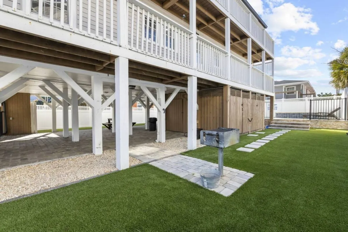 The image captures an outdoor grill setup beneath a deck, with a grassy area and stone pathway nearby. The grill area is ideal for barbecuing, socializing, and enjoying meals with family and friends in a relaxed, sunny setting.