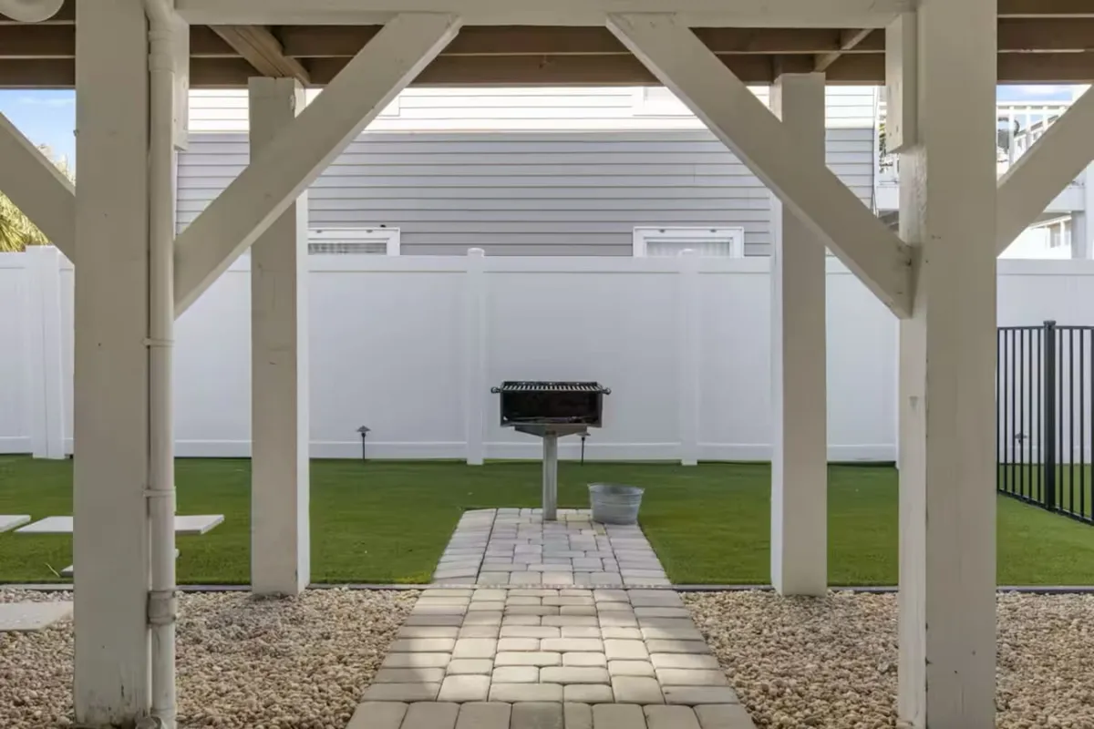 A relaxed exterior scene showing a grill, pathway, green lawn, and fenced yard. The setting is designed for outdoor dining and social gatherings, with ample space for barbecues and celebrations under the sky.