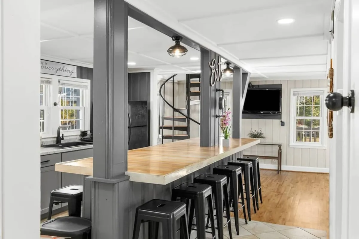 A lively, contemporary interior featuring a long wooden bar counter lined with black bar stools, a spiral staircase adding architectural interest, and a wall-mounted TV. The space exudes a sense of social energy, perfect for casual dining and entertaining guests.