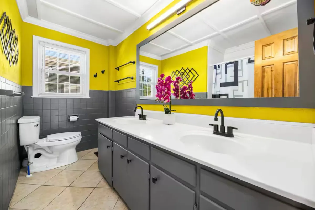 A vibrant bathroom featuring a double vanity with sinks, bright yellow walls, a decorative mirror, and fresh purple orchids. The mood is uplifting and refreshing, ideal for starting the day or unwinding at night.