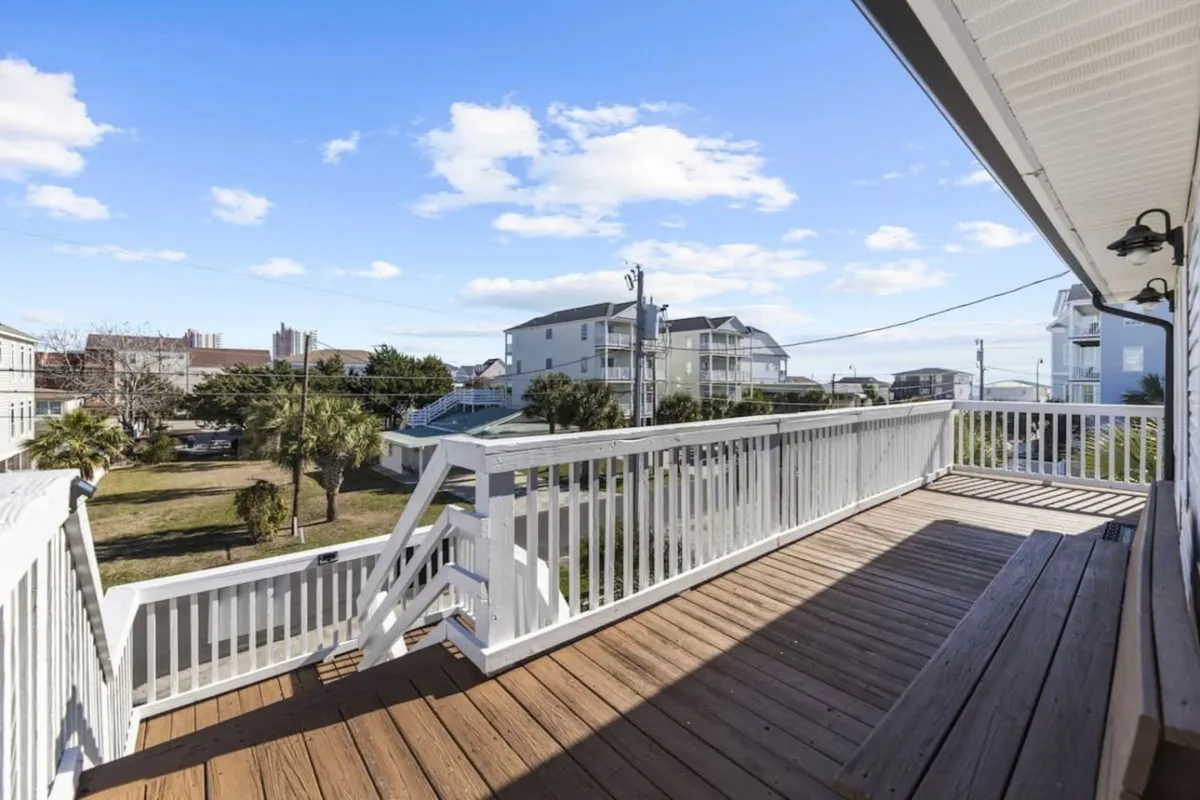 A spacious wooden deck with white railings overlooks neighboring houses and lush green palm trees. The deck suggests opportunities for relaxing, sunbathing, or enjoying coffee while taking in the surrounding neighborhood and preparing for a day of exploration.